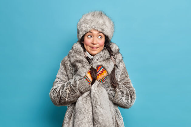 模特体贴的因纽特人妇女 戴着灰色皮帽和外套 戴着针织手套 穿着温暖的冬装 隔着蓝色的工作室墙壁时尚正面微笑