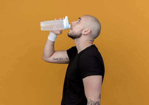 穿站在侧视图年轻的运动男子戴着腕带喝水从水瓶隔离在橙色的墙壁男人轮廓观点