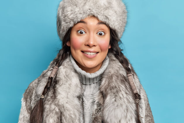 霜冻一位留着两条辫子 身穿毛灰色外套的亚洲妇女的特写镜头 微笑着愉快地生活在隔离在蓝色墙壁上的北极地区民族手套头饰