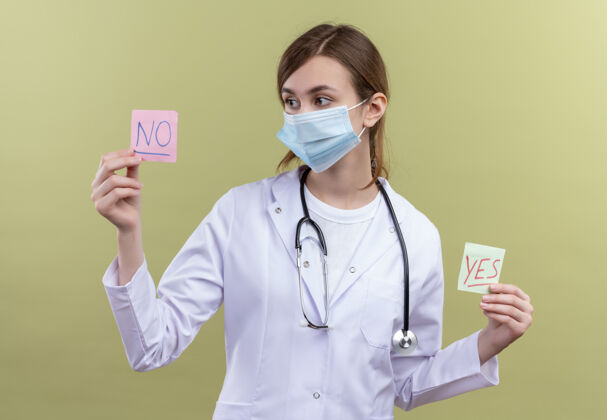 口罩年轻的女医生穿着医用长袍 戴着口罩和听诊器 拿着是和否的纸条 看着隔离的绿墙上没有纸条长袍便条医生