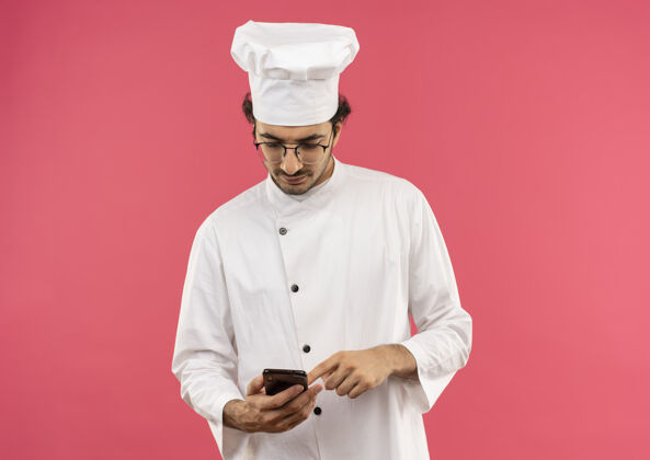 手持想着年轻的男厨师穿着厨师制服 戴着眼镜 拿着手机看着隔离在粉红色墙上的墙佩戴电话烹饪