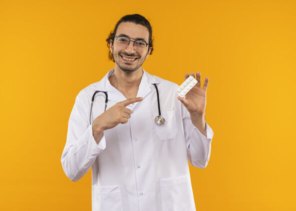 年轻面带微笑的年轻医生 戴着医用眼镜 穿着医用长袍 手持听诊器 指着隔离的黄色墙壁上的药片 还有复印空间医生听诊器点