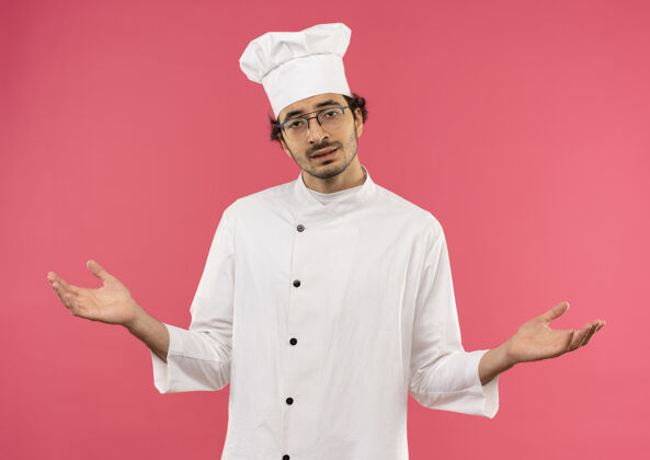 烹饪年轻的男厨师穿着厨师制服 戴着眼镜 双手分开放在粉红色的墙上男性不愉快手