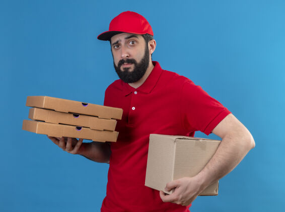男人困惑的年轻英俊的白人送货员穿着红色制服 戴着帽子 拿着纸箱和披萨包裹 隔离在蓝色的上面纸箱盒子送货