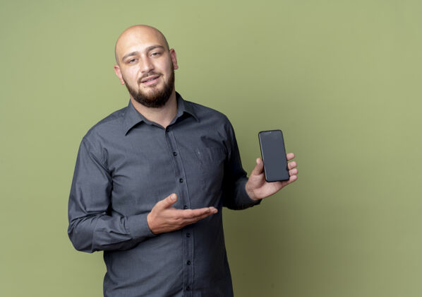 指向自信的年轻秃头呼叫中心的男子显示 手持和指着手机孤立的橄榄绿与复制空间橄榄手电话