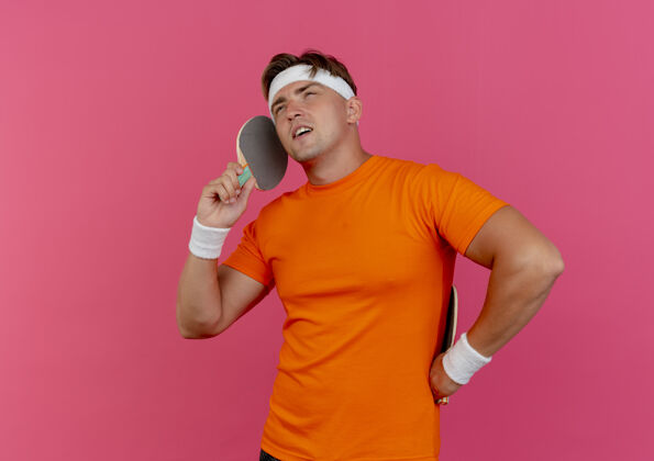 自信自信的年轻帅气的运动型男人戴着头带和腕带 拿着乒乓球拍 假装在电话里说话 粉红色的地板上有一个复制空间穿太空球拍