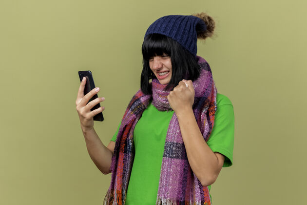 橄榄快乐的年轻病高加索女孩戴着冬天的帽子和围巾拿着看手机拿着餐巾做是的手势孤立在橄榄绿的背景与复制空间手势快乐年轻