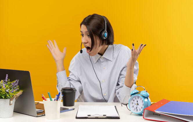 耳机印象深刻的年轻呼叫中心的女孩戴着耳机坐在办公桌旁举手 看着笔记本电脑上孤立的橙色坐着笔记本电脑女孩