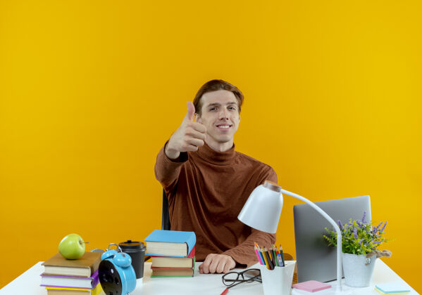 工具微笑的年轻学生男孩坐在书桌旁 手里拿着学习工具 他的大拇指孤立地竖在黄色的墙上学校男孩学生