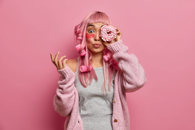 粉色有趣的亚洲粉红头发女人头上有滚轴 用美味的甜甜圈遮住眼睛 戴上胶原蛋白贴片来减少皱纹刷新美容贴片