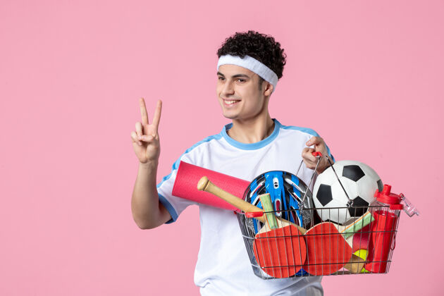 完整正面图穿着运动服的年轻男子 带着装满运动用品的篮子 粉色的墙壁篮子成人肖像