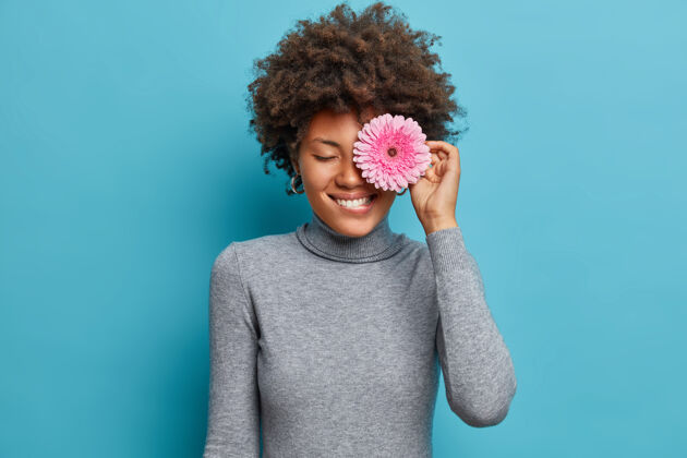 姿势美丽的非裔美国妇女的肖像覆盖着玫瑰色非洲菊的眼睛 咬着嘴唇 积极地微笑 喜欢花 穿着休闲的高领毛衣快乐模特牙关