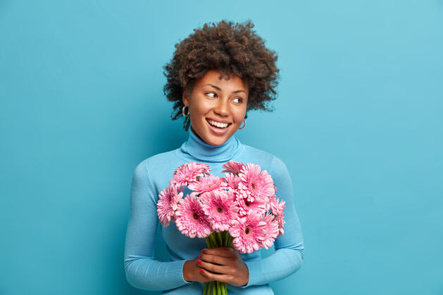 女性快乐的非洲裔美国女人庆祝她特殊的日子 捧着一束粉红色的花 崇拜非洲菊 笑容灿烂 穿着高领毛衣 人微笑年轻