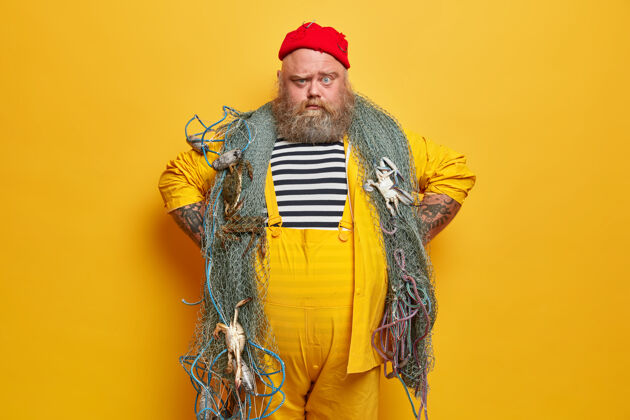 超重严肃的渔夫表情自信 双手放在臀部 穿着条纹水手衬衫和黄色工作服 准备用网捕鱼姿势职业人类