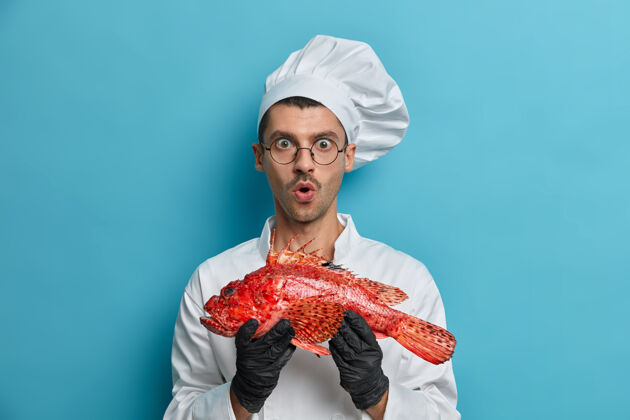 拼图惊讶的男人拿着生的红鱼 张着嘴 准备烤或煮鲈鱼 穿着制服 戴着橡胶手套室内成人食物