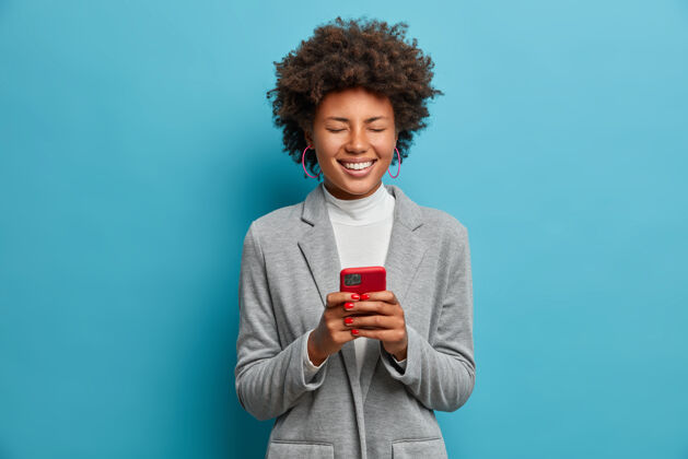 时尚开朗的美国黑人女博主在网上发布图片 通过电话工作 在网上看搞笑视频 开心地闭上眼睛 穿着灰色夹克 卷发民族女士