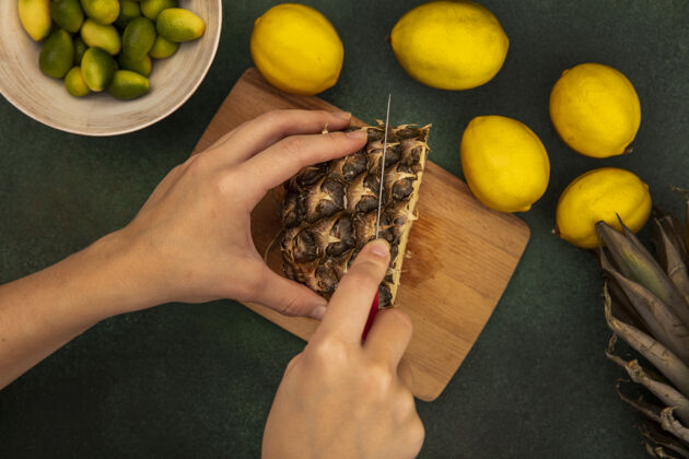 甜点俯视图中的女性手切割新鲜菠萝在木制厨房板上用刀与金盏花在一个碗与柠檬隔离在一个绿色的墙壁特写板菠萝