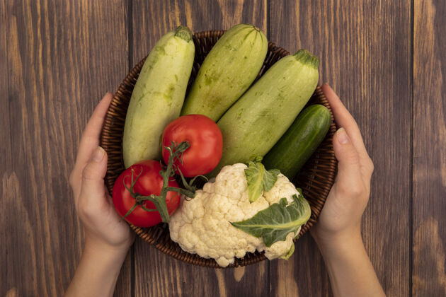 农业顶视图：女性手拿着一桶新鲜蔬菜 比如西葫芦 西红柿 黄瓜和菜花放在木墙上新鲜蔬菜黄瓜
