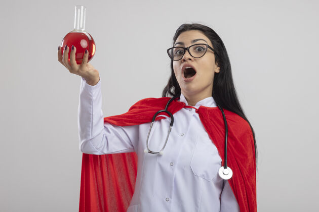 衣服印象深刻的年轻女超人戴着眼镜和听诊器举起装有红色液体的化学瓶向上望着前面隔离在白色墙上斗篷表情瓶子