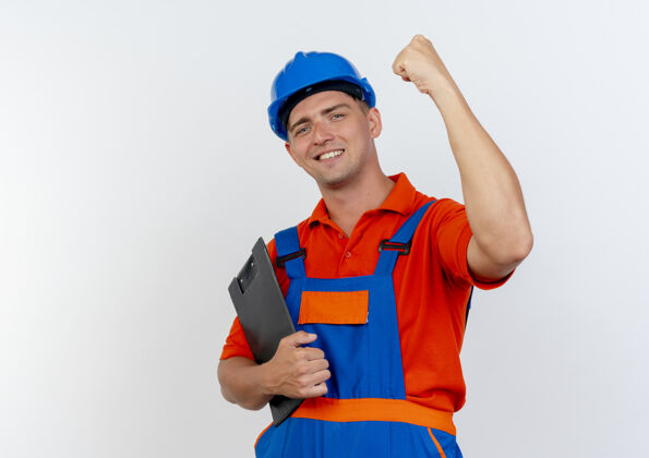 安全面带微笑的年轻男性建筑工人穿着制服 戴着安全帽 拿着写字板 在白色地板上做着有力的手势手势微笑剪贴板