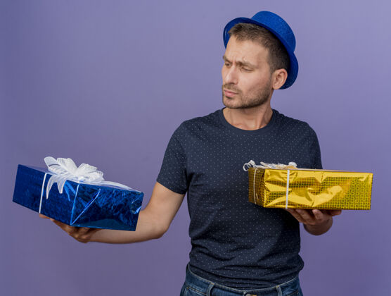 帅哥困惑的帅哥戴着蓝色的帽子 拿着礼物盒 看着紫色的墙壁上孤立感觉持有市民