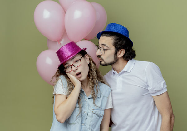 橄榄色一对戴着粉蓝色帽子的年轻夫妇站在气球前 女孩把手放在脸颊上绿色帽子亲吻