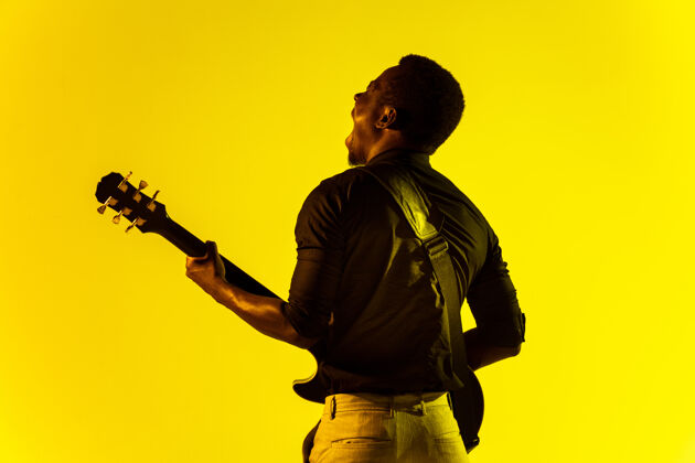 旋律年轻的非洲裔美国音乐家在霓虹灯下像摇滚明星一样在黄色背景下弹吉他节日霓虹灯衬衫