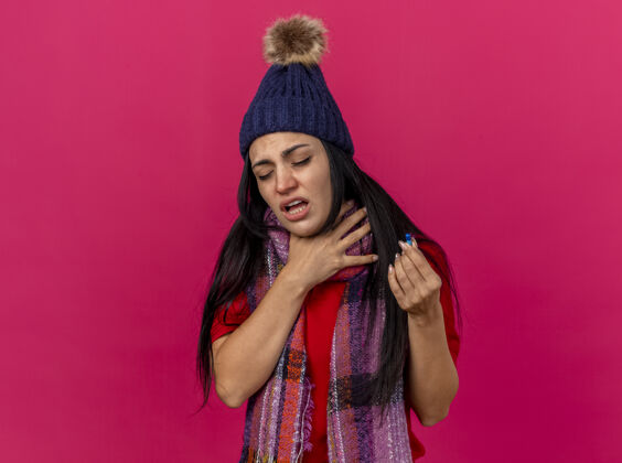 人一个戴着冬日帽子 戴着围巾 拿着胶囊 眼睛闭着 喉咙被隔离在粉红色墙壁上的年轻病女人衣服感觉脸