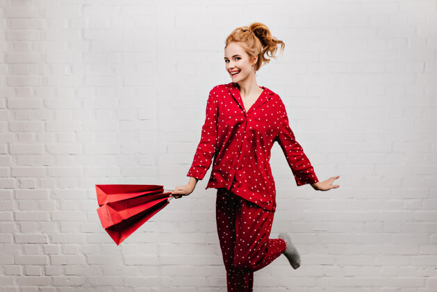 年轻纸袋激发年轻女子的灵感 表达活力室内照片：波浪发女士穿着红色睡衣 手持新年礼物金发服装身体