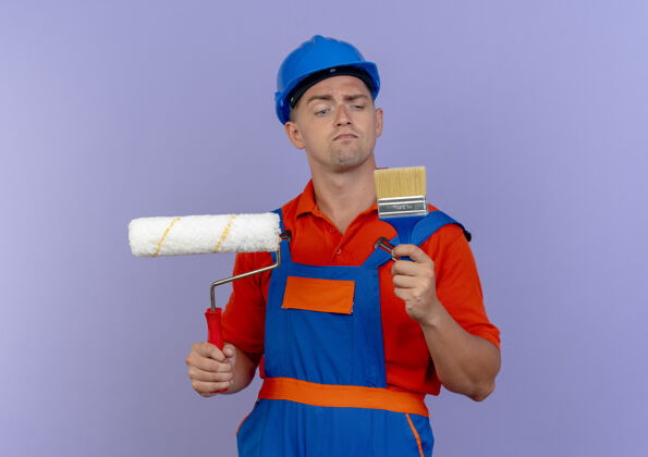 头盔想着年轻的男建筑工人穿着制服 戴着安全帽 手里拿着油漆辊 看着油漆刷上的紫色制服油漆思考