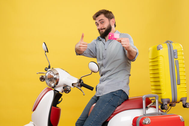 汽车旅游概念的正面视图 一个快乐微笑的年轻人坐在摩托车上 背着手提箱 手里拿着黄色的银行卡摩托车持有微笑