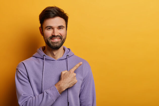年轻人正面胡须男人的横拍显示在空白处 开心地微笑 展示漂亮的广告 穿着紫色的运动衫 隔离在黄色的墙壁上人物和宣传概念促销表演表情