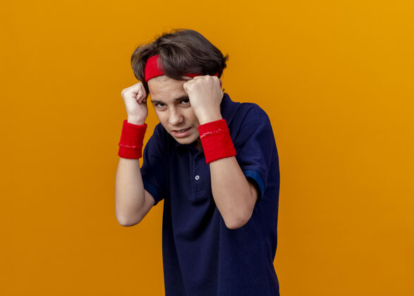 戴年轻帅气的运动男孩 戴着头带和护腕 戴着牙套 拳头紧靠着脸 看着橙色墙壁上孤立的前方人保持靠近