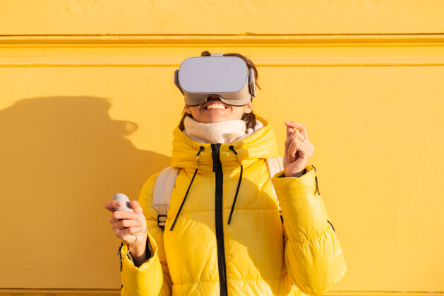 模拟一个戴着虚拟现实眼镜的女人的肖像 在冬天的阳光下 穿着暖和的衣服 靠在黄色的墙上显示微笑眼镜