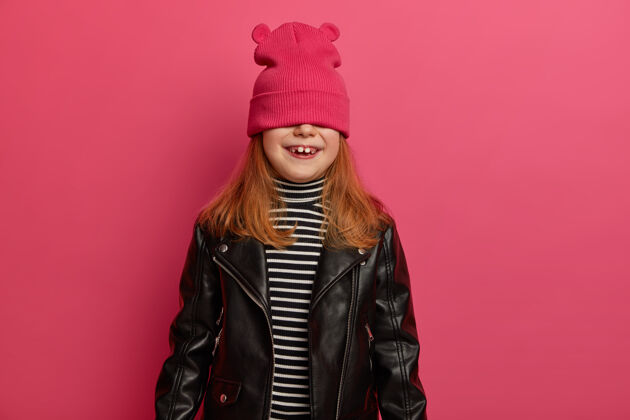 孩子可爱的红发女孩玩捉迷藏游戏 用积极的情绪等待惊喜 用粉色帽子遮住眼睛 穿条纹套头衫和皮夹克 玩得很开心 在室内摆姿势小高兴高兴