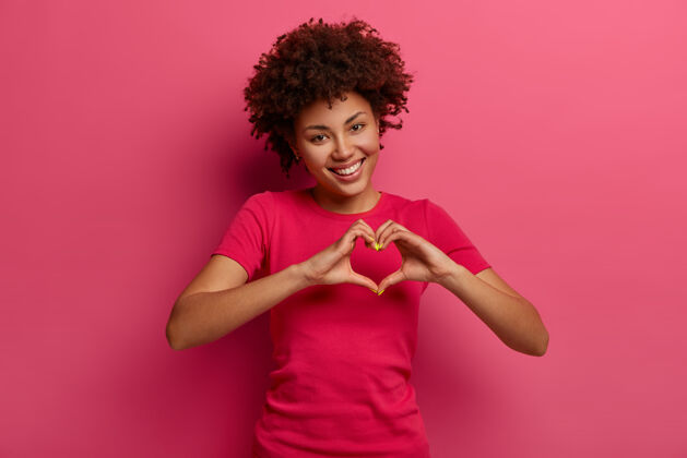 女人漂亮的卷发的非洲裔美国女人承认爱 做心形手势 展示她的真情 有快乐的表情 穿着休闲的红色t恤 在粉红色的墙上摆姿势关系概念罗西显示手势