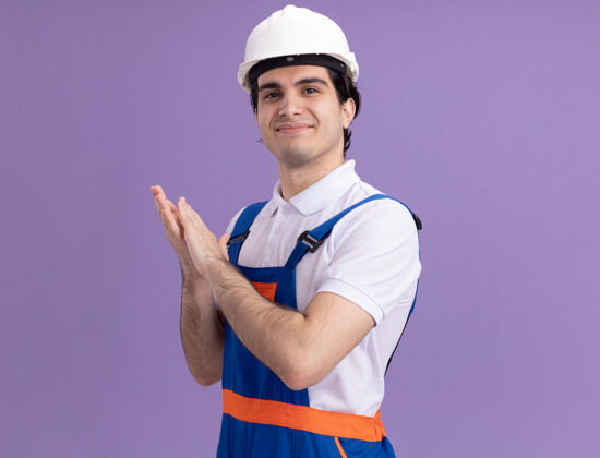 安全年轻的建筑工人穿着建筑制服 戴着安全帽 站在紫色的墙上 微笑着自信地鼓掌制服自信男人