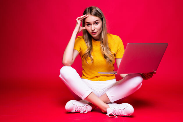 坐着快乐的年轻卷发美女盘腿坐在地板上 在红墙上用笔记本电脑人舒适成功