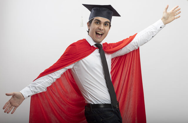 手快乐的年轻超级英雄看着戴着领带和毕业帽的相机 双手分开 背景是白色的传播领带相机