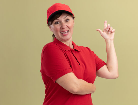 伟大身着红色制服 头戴鸭舌帽的中年女送货员站在绿色的墙边 微笑着自信地展示着有新创意的食指目录自信分娩