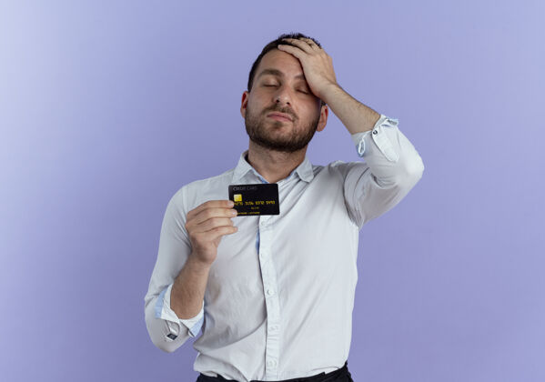 成人累了的帅哥把手放在额头上拿着信用卡隔离在紫色的墙上英俊男人人