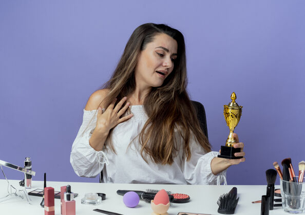 优胜者惊讶的美丽女孩坐在桌旁 用化妆工具把手放在胸前 看着紫色墙上孤立的优胜者杯胸部杯子坐着