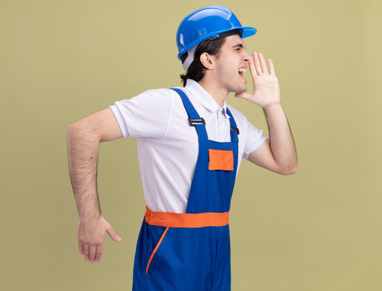 呼叫身穿施工制服 戴安全帽的年轻建筑工人站在绿墙旁 用手喊或叫人靠近制服头盔
