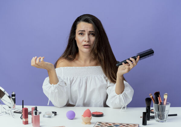 桌子悲伤美丽的女孩坐在桌子旁 拿着化妆工具 把梳子孤立在紫色的墙上美女工具女人