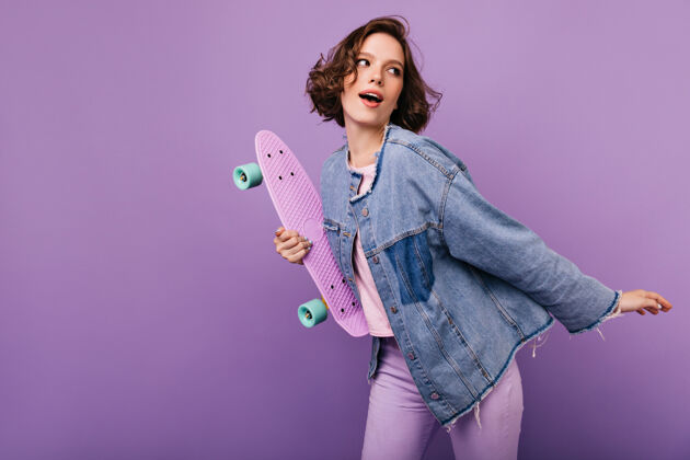 欢呼穿着超大号夹克跳舞的迷人的年轻女士梦幻卷曲的女孩和滑板的肖像配件女人紫色