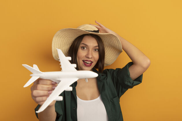 姿势快乐美丽的高加索女人戴着沙滩帽拿着橙色的飞机模型漂亮黑发沙滩