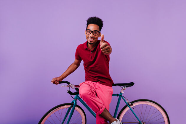 时尚灵感来自戴眼镜的非洲男人 大拇指朝上摆姿势自信的黑人坐在自行车上微笑着成人非洲男孩