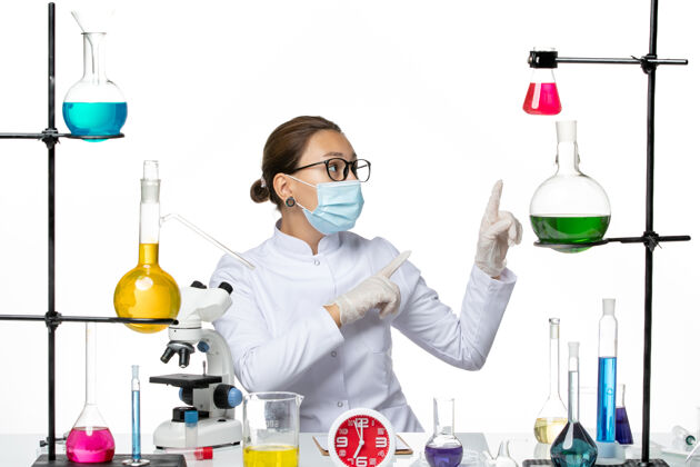 专业前视图：穿着医疗服的女化学家 戴着面罩 坐在白色办公桌上 拿着溶液 化学病毒 病毒 喷溅科学解决方案化学