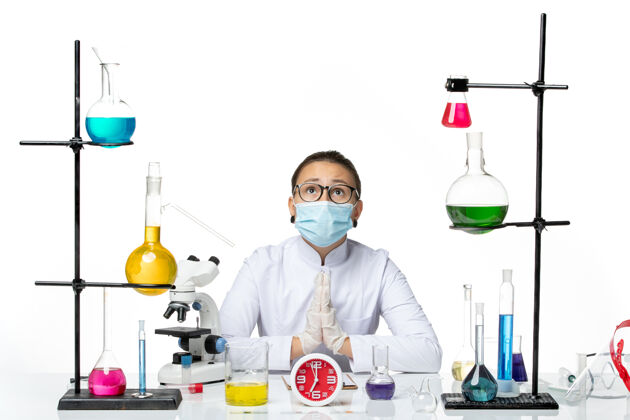 查看前视图：穿着医疗服的女化学家 戴着面罩 坐在白色背景上祈祷 病毒实验室的化学成分会喷溅解决方案科学专业