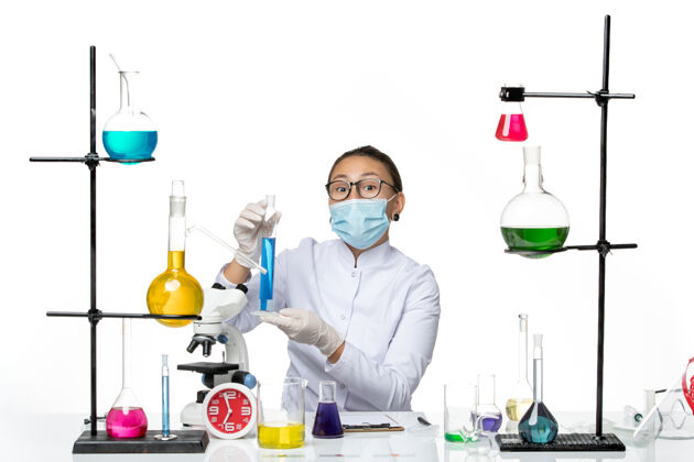 医学正面图：穿着医疗服的女化学家戴着面罩 白色桌子上拿着蓝色溶液 溅起病毒化学病毒前面保持喷溅
